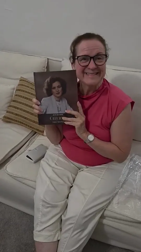 Cheryl on her couch, holding up her finished book, with her own portrait on the cover.